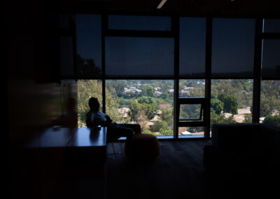 a person is silhouetted while peeking through the blinds of a 4th story building looking at beautiful trees.