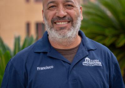 headshot of an older man, outlined by building and palm trees behind him.